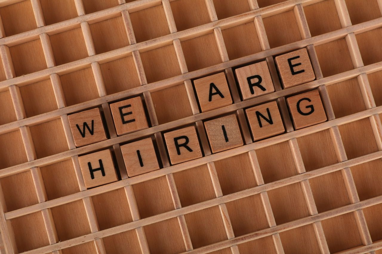 About Wooden letter tiles arranged to spell 'We Are Hiring' on a wooden grid background.