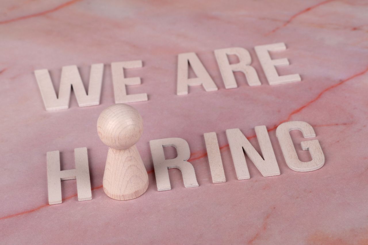 Home Wooden letters spelling 'We Are Hiring' on a pink marble surface with a pawn piece.